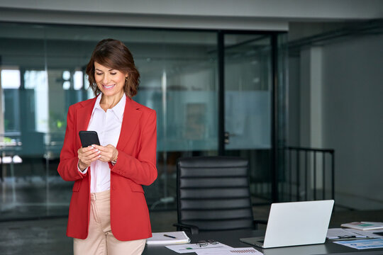 Smiling Latin Hispanic middle-aged female businesswoman holding smartphone trading, working online at office workplace computer. Mature European business woman manager ceo using cell phone. Copy space - Powered by Adobe