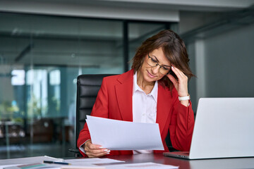 Confused mature trader businesswoman looking disappointed at loan tax paper document using laptop...