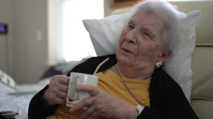 An elderly woman sits comfortably in a home healthcare facility, sipping a warm drink and enjoying her surroundings in tranquility.