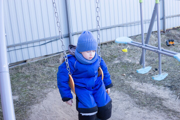 Kid in blue jumpsuit and hat swinging gently on a outdoor swing in a backyard. Childhood activity and relaxation concept for family play and park fun.
