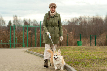 Woman walks with her dog welsh corgi in park. Stylish lady enjoys playtime whith her pet. Autumn season. Fall time.