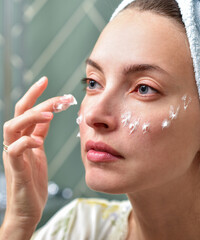 Beautiful Young Woman applying facial moisturizing cream in bathroom.Home interior. Skincare concept