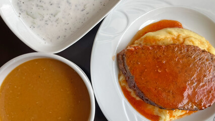 Served on a plate; a slice of roast beef on mashed potatoes, with soup and yogurt dip on the side.
