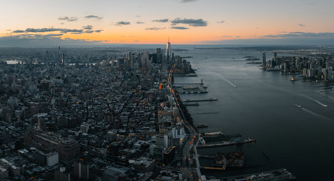 Aerial view of a sprawling cityscape where the Hudson River reflects the warm glow of the setting sun, illuminating the iconic One World Trade Center, New York, New York, United States.