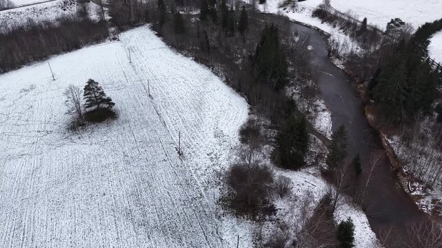 Aerial view of serene winter landscape featuring snow-covered fields, winding river, and scattered trees, showcasing gradual camera zoom out revealing expansive scenery