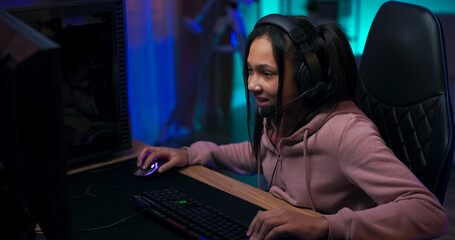 Teenage girl passionate about the virtual world plays shooters on the computer, young girl excitement vigorously clicks fingers on the keyboard, has headphones, backlit accessories blue room lighting.