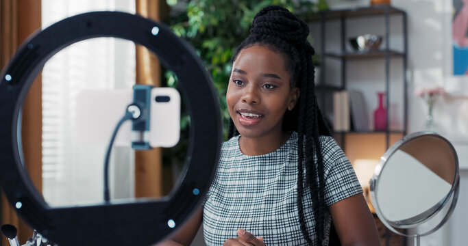 Influencer smiles at camera from apartment, makes video for social media about makeup products, sitting by mirror with ring light and phone tripod, working remotely online.