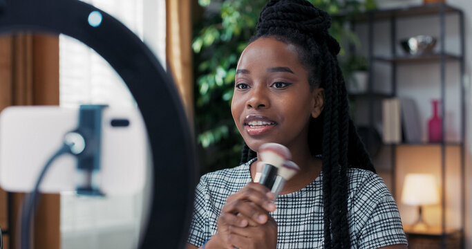 A smiling beauty creator records a video at home using a ring light and phone. She presents her favorite makeup brushes for foundation and bronzer, highlighting soft, fluffy bristles.