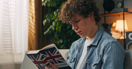 A young male student prepares for an English exam in his dorm, reading carefully from a language learning textbook.