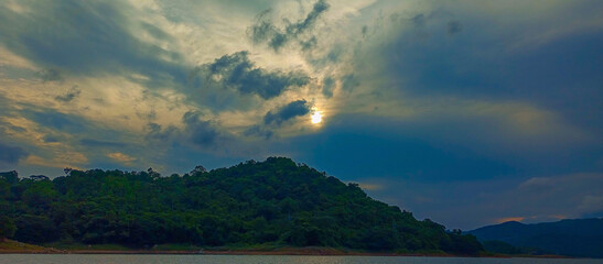 The sun attempts to break through dark, dramatic clouds above a lush, green, densely forested hill rising from the water's edge at dusk.