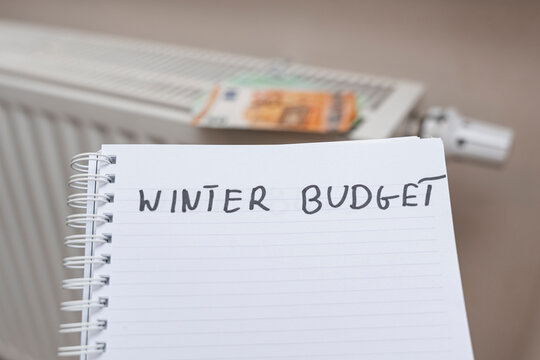 Handwritten note titled WINTER BUDGET on a blank notebook page, with a background featuring a radiator and banknotes, symbolizing financial planning and winter expenses. - Powered by Adobe