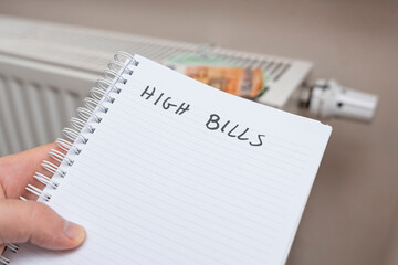 A handwritten note reading HIGH BILLS on a spiral notebook held by a person next to a radiator. This image symbolizes financial stress and household expenses.