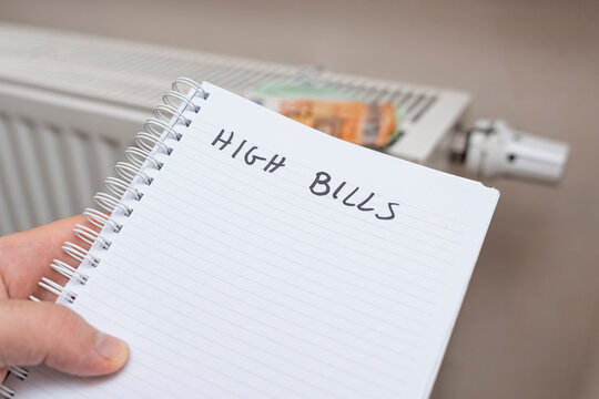 A handwritten note reading HIGH BILLS on a spiral notebook held by a person next to a radiator. This image symbolizes financial stress and household expenses.