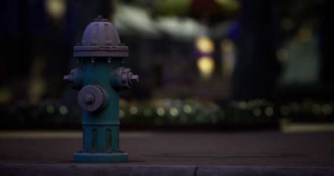 A bright blue fire hydrant is positioned on a walkway in an urban park as night descends. Surrounding lights create a magical atmosphere, highlighting the hydrants vivid color.
