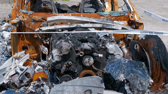 Burned car wreckage under police line. Burned car wreckage stands behind a police line, showing extensive damage from fire and potential vandalism, possibly after an accident or arson