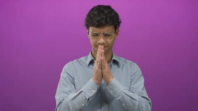 Man wearing glasses pressing hands together in purple studio for prayer pose; serenity calm reflection mindfulness.