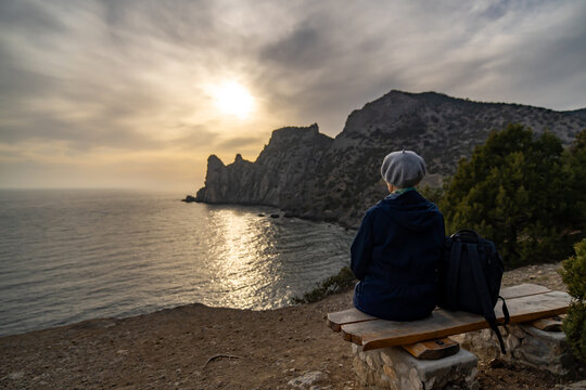 Traveler sunset seascape, person sitting on cliff overlooking sea and large mountains