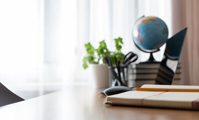 Notebook and pencil on a desk with blurred home office elements in the background &mdash; laptop, mouse, books, globe and plant. Cozy remote work, studying or teaching from home concept.