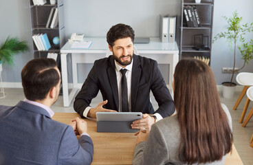 Male financial advisor consulting young couple about insurance sitting at the desk on workplace in office using tablet. Man and woman talking with realtor or business broker going to sign a contract.