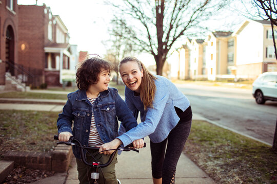 Adult mother and young son laughing while learning to bike on sidewalk