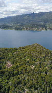 Aerial view of vibrant green forest reaching the edge of a serene blue lake against the backdrop of distant mountains, San Carlos de Bariloche, Rio Negro Province, Argentina.