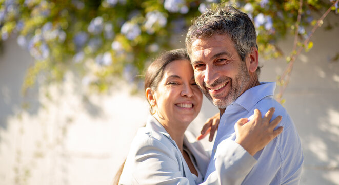 Happy couple embracing and smiling under sunny day in front of a white wall with flowers
