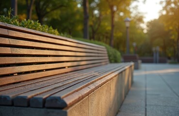 Empty modern wooden bench waits in sunlit park. Concrete base supports seat, paving tiles nearby. Trees and green bushes form background blur.