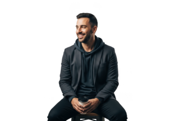 A smiling man with dark hair and a beard wearing a dark hoodie and pants sits with his hands clasped isolated on transparent background