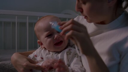 Mother gently cleans baby's nose with a nasal aspirator while seated in a cozy nursery, showcasing the tender interaction, camera pans to capture the moment