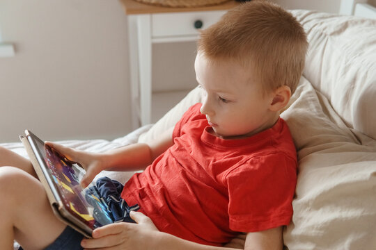 Little boy using a digital tablet while relaxing on a bed at home. Kid playing mobile game or watching video. Concept of child screen time. - Powered by Adobe