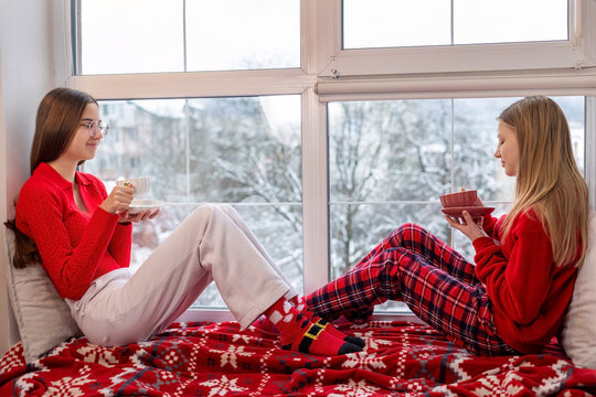 Teen girls relaxing by window with hot drinks in winter. Two teenage girls in cozy clothes sitting on a window seat with hot drinks, enjoying a snowy winter day and holiday vibes. - Powered by Adobe