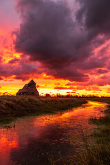 The iconic St Thomas &agrave; Becket Church on Fairfield, Kent stands silhouetted against a blazing sunrise, dramatic clouds rolling across the Romney Marsh landscape.