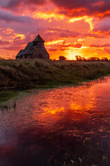 The iconic St Thomas &agrave; Becket Church on Fairfield, Kent stands silhouetted against a blazing sunrise, dramatic clouds rolling across the Romney Marsh landscape.
