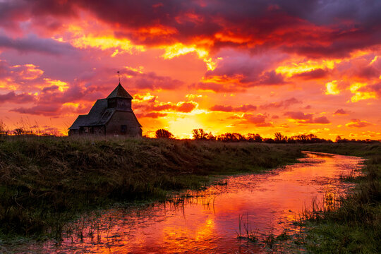 The iconic St Thomas &agrave; Becket Church on Fairfield, Kent stands silhouetted against a blazing sunrise, dramatic clouds rolling across the Romney Marsh landscape.