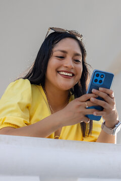 Mujer sonriente con gafas de sol sobre la cabeza, de blusa amarilla, sonriendo mientras mira su tel&eacute;fono m&oacute;vil azul.