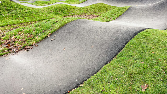 Curved asphalt pump track for cycling and wheeled sports, surrounded by vibrant green grass in a modern park