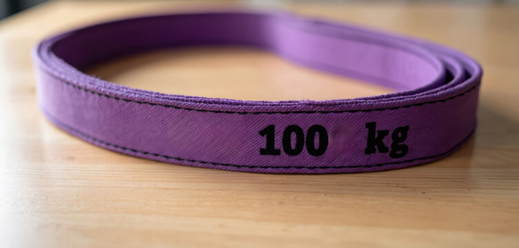 Purple lifting sling marked 100 kg rests on wood surface. Heavy duty industrial strap for cargo, safe load lifting. Rigging equipment, construction safety tool.