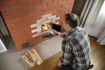 Naklejka premium Caucasian adult man adding large log into burning brick stove while warming indoor room during cold season. Cold season. Winter Firewood Routine Preparing and Lighting Wood Stove