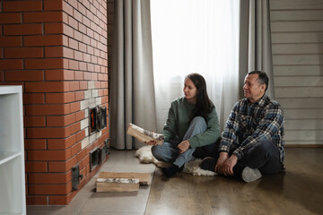 Naklejka premium Adding firewood to heat stove by caucasian man and woman sitting together on floor in warm indoor rustic room with natural light. Winter Firewood Routine Preparing and Lighting a Wood Stove