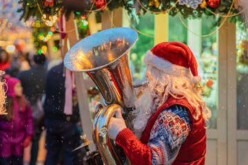 Santa Claus playing tuba at outdoor Christmas market with festive decorations and falling snow....