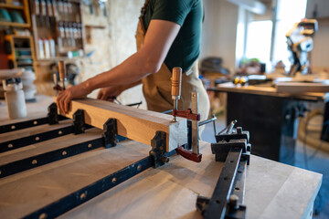 Man carpenter working in carpentry workshop, fixing wooden blocks at table with special clamps for compression and gluing, working process on precise alignment condition, creating new furniture item.