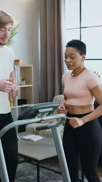 Home workout concept where beautiful active sporty african american woman supporting her muscular bearded freind which jogging on running track,she setting the speed on machine's control panel