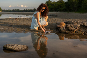 Woman playing with paper boat in water puddle on sandy beach at sunset, reflecting childhood