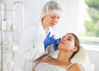 Obraz premium Female doctor inserts filler into patients lip area to correct volume and shape. Permanent makeup