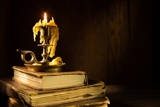 A candle in a brass holder is melting, resting atop a stack of old, worn books. Candle on vintage books creates a warm atmosphere in dimly lit room