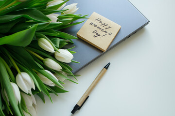 Sticky note with Women's day handwritten congratulations and white tulips bouquet laying on the laptop at white office table. Greetings for women at workplace.