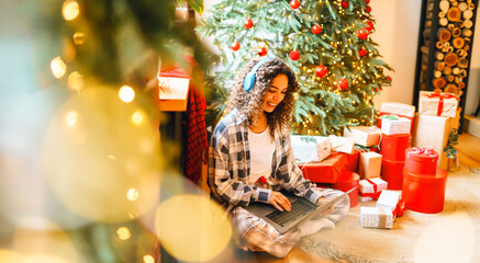 Portrait of woman in pajamas and Santa hat with laptop surrounded by holiday gifts in cozy room. Happy woman works on laptop near decorated Christmas tree. Holiday concept, freelance.