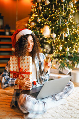 Portrait of woman in pajamas and Santa hat with laptop surrounded by holiday gifts in cozy room....