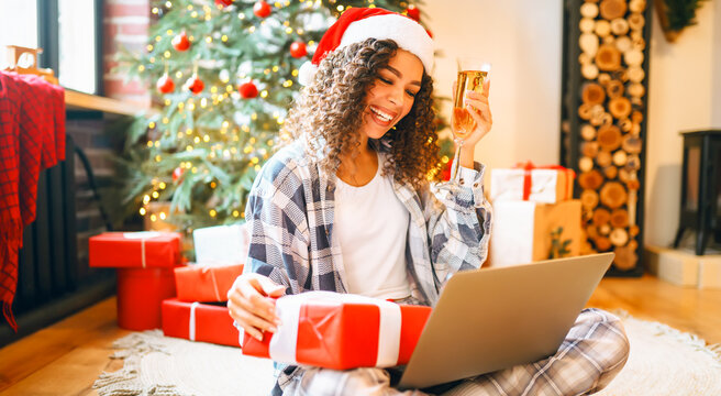 Portrait of woman in pajamas and Santa hat with laptop surrounded by holiday gifts in cozy room. Happy woman works on laptop near decorated Christmas tree. Holiday concept, freelance. - Powered by Adobe