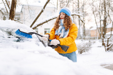 Fototapeta premium A woman in bright jacket clears snow from car windshield in morning in snowy area. Female driver clears snow from car window on sunny winter day. Concepts of transportation, cleaning, and seasonality.